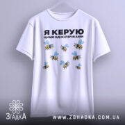 Футболка керую бджілочками, білого кольору, підвішена на вішалці, фон світло-сірий, принт з бджолами та написом "Я КЕРУЮ ЦИМИ БДЖІЛОЧКАМИ" в чорному кольорі.