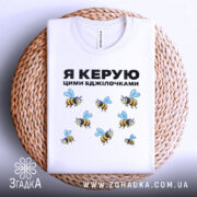 Футболка керую бджілочками, білого кольору, складена на плетеній кошику, фон світло-сірий, принт з бджолами та написом "Я КЕРУЮ ЦИМИ БДЖІЛОЧКАМИ" в чорному кольорі.