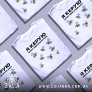 Футболка керую бджілочками, білого кольору, зображена на манекені з правого ракурсу, фон світло-сірий, принт з бджолами та написом "Я КЕРУЮ ЦИМИ БДЖІЛОЧКАМИ" в чорному кольорі.