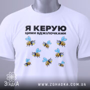 Футболка керую бджілочками, білого кольору, складена на поверхні, фон світло-сірий, принт з бджолами та написом "Я КЕРУЮ ЦИМИ БДЖІЛОЧКАМИ" в чорному кольорі, з зеленим ярликом.