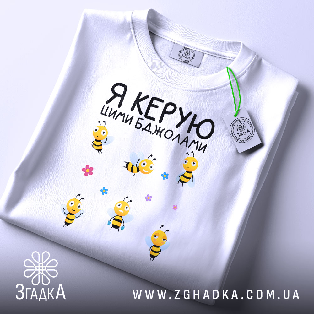 Футболка керую бджолами білого кольору, складена на столі, фон світло-сірий, принт з жовтими бджолами та квітами, на горловині видна етикетка.