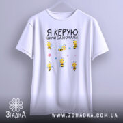 Футболка керую бджолами білого кольору, підвішена на вішалці, фон світло-сірий, принт з жовтими бджолами та квітами розташований по центру.