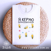 Футболка керую бджолами білого кольору, складена на плетеному кошику, фон білий, принт з жовтими бджолами та квітами, видна етикетка на горловині.