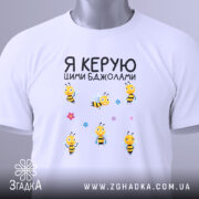 Футболка керую бджолами білого кольору, складена на столі, фон світло-сірий, принт з жовтими бджолами та квітами, на горловині видна етикетка.