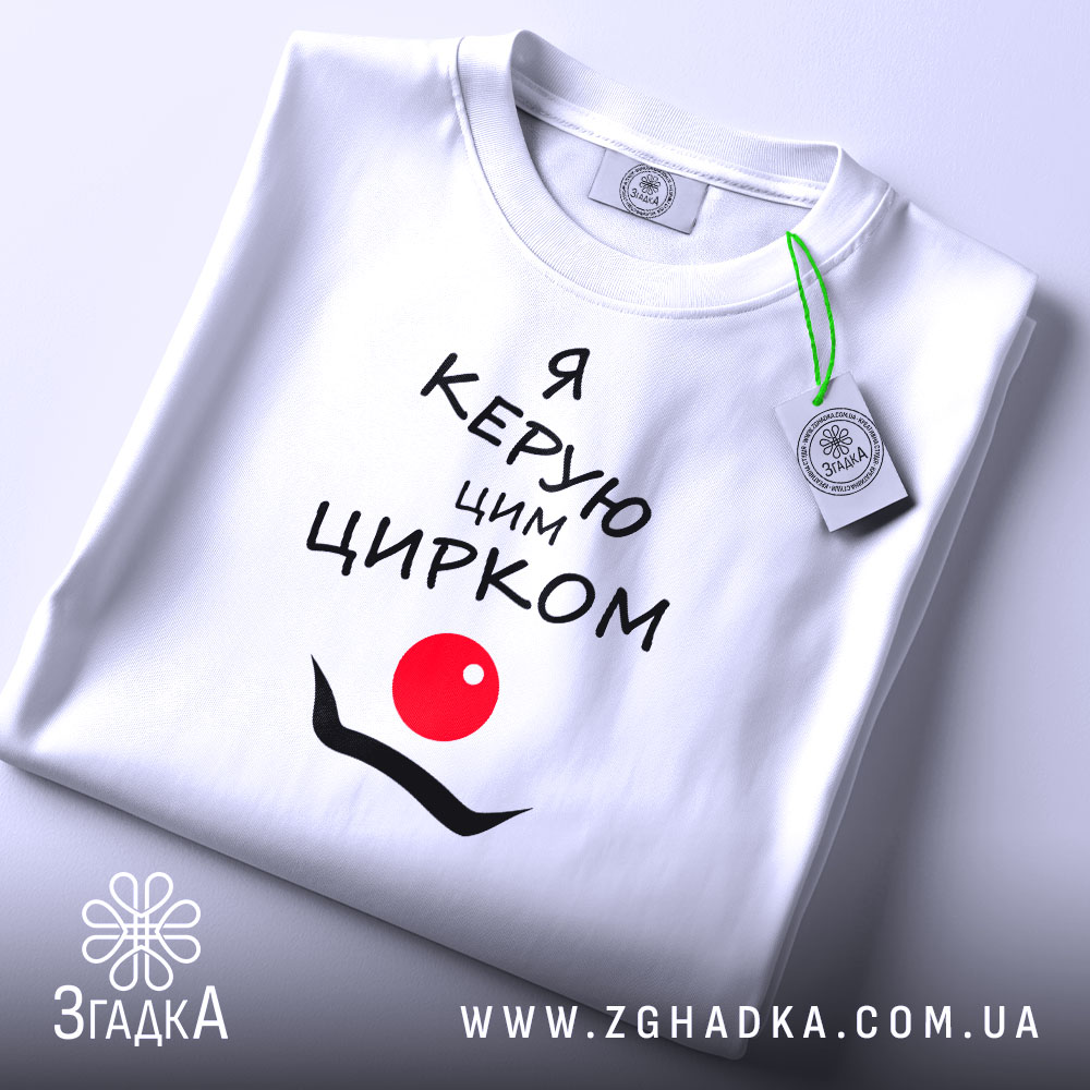 Футболка керую цирком білого кольору, складена на столі, з принтом чорного кольору, що містить текст "Я керую цим цирком" та червоний круг, з зеленим ярликом на комірці, на фоні світло-сірого кольору.