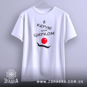 Футболка керую цирком білого кольору, підвішена на вішалці, з принтом чорного кольору, що містить текст "Я керую цим цирком" та червоний круг, на фоні світло-сірого градієнту.