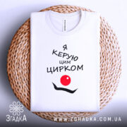 Футболка керую цирком білого кольору, складена на плетеній кошику, з принтом чорного кольору, що містить текст "Я керую цим цирком" та червоний круг, на фоні світло-сірого кольору.