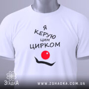 Футболка керую цирком білого кольору, складена на столі, з принтом чорного кольору, що містить текст "Я керую цим цирком" та червоний круг, з зеленим ярликом на комірці, на фоні світло-сірого кольору.