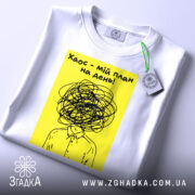 Футболка хаос мій план, біла, розкладена на поверхні, фон світло-сірий, принт з жовтим фоном і чорним малюнком, що зображає людину з заплутаним волоссям, з етикеткою на рукаві.