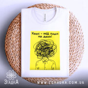 Футболка хаос мій план, біла, розкладена на плетеній кошику, фон білий, принт з жовтим фоном і чорним малюнком, що зображає людину з заплутаним волоссям.