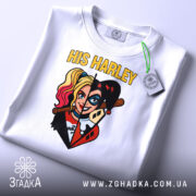 Футболка Харлі Квін білого кольору, складена на столі, з видимим етикеткою, фон світло-сірий. Принт зображає Харлі Квін з двома різними стилями, з написом 'HIS HARLEY' у верхній частині.
