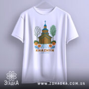 Футболка Хажин білого кольору, підвішена на вішалці, фон світло-сірий, принт з дерев'яною церквою, річкою та квітами, текст 'KHAZHYN UKRAINE' внизу.