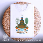 Футболка Хажин білого кольору, зображена на плетеній кошику, фон світлий, принт з дерев'яною церквою, річкою та квітами, текст 'KHAZHYN UKRAINE' внизу, видна етикетка на комірці.