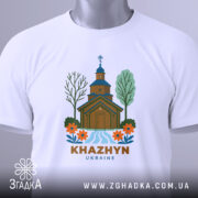 Футболка Хажин білого кольору, зображена на столі, ракурс зверху, фон світлий, принт з дерев'яною церквою, річкою та квітами, текст 'KHAZHYN UKRAINE' внизу, на комірці видна етикетка.