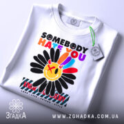 Футболка хейт квітка стиль, біла футболка на столі, з принтом квітки та текстом, вид з верхнього ракурсу, на футболці є етикетка з зеленим шнурком, фон світло-сірий.