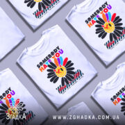 Футболка хейт квітка стиль, біла футболка на манекені, з принтом у вигляді квітки з усміхненим обличчям, кольори принта яскраві: червоний, жовтий, синій, чорний, фон світло-сірий.