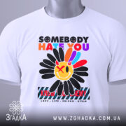 Футболка хейт квітка стиль, біла футболка на столі, з принтом квітки та текстом, вид з верхнього ракурсу, на футболці є етикетка з зеленим шнурком, фон світло-сірий.