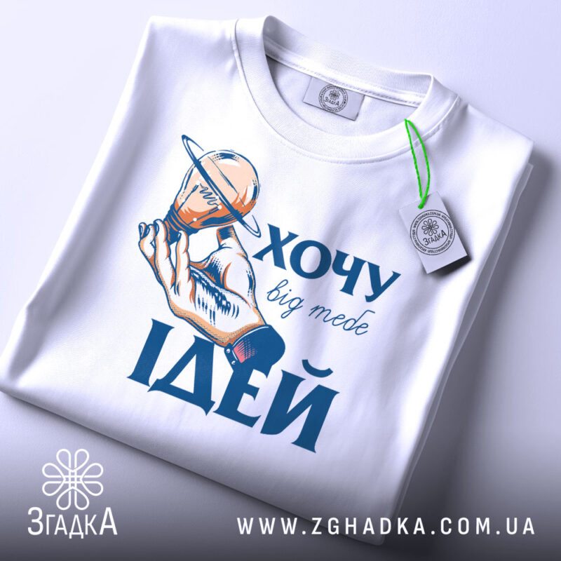 Футболка хочу ідей білого кольору, складена на поверхні, фон світло-блакитний, принт з рукою, що тримає лампочку з кільцем, текст "ХОЧУ big mebe ІДЕЙ" у синьому кольорі, на футболці є етикетка з зеленим шнурком.