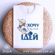 Футболка хочу ідей білого кольору, складена на плетеній кошику, фон світло-блакитний, принт з рукою, що тримає лампочку з кільцем, текст "ХОЧУ big mebe ІДЕЙ" у синьому кольорі.
