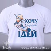 Футболка хочу ідей білого кольору, складена на поверхні, фон світло-блакитний, принт з рукою, що тримає лампочку з кільцем, текст "ХОЧУ big mebe ІДЕЙ" у синьому кольорі, на футболці є етикетка з зеленим шнурком.