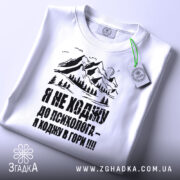 Футболка ходжу в гори, білого кольору, складена на поверхні, фон світло-сірий, принт з горами та текстом чорного кольору, з зеленим ярликом.