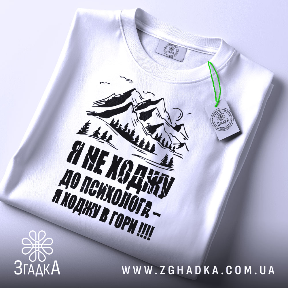 Футболка ходжу в гори, білого кольору, складена на поверхні, фон світло-сірий, принт з горами та текстом чорного кольору, з зеленим ярликом.