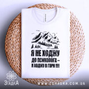 Футболка ходжу в гори, білого кольору, складена на плетеній основі, фон світло-сірий, принт з горами та текстом чорного кольору.