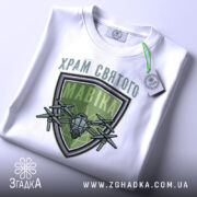 Футболка храм святого мавіка, біла, складена на поверхні, фон світло-сірий, принт з зеленим щитом і дронами, на футболці є етикетка.