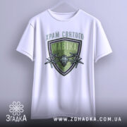 Футболка храм святого мавіка, біла, підвішена на вішалці, фон світло-сірий, принт з зеленим щитом і дронами, текст зверху на українській мові.