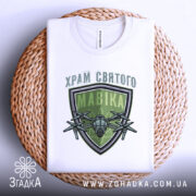 Футболка храм святого мавіка, біла, складена на плетеній кошику, фон білий, принт з зеленим щитом і дронами, текст зверху на українській мові.