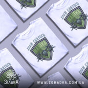 Футболка храм святого мавіка, біла, зображена на манекені, фон світло-сірий, принт з зеленим щитом і дронами, текст зверху на українській мові.