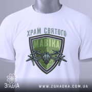 Футболка храм святого мавіка, біла, складена на поверхні, фон світло-сірий, принт з зеленим щитом і дронами, на футболці є етикетка.