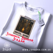 Футболка хвилина мовчання. Біла футболка, складена на поверхні, з видимим принтом на грудях. На принті зображені солдати біля свічки, текст "ПАМ'ЯТЬ - ЗБРОЯ!" та червоний напис "Зупинись. Вшануй. Згадай.". Видно етикетку з зеленим шнурком.