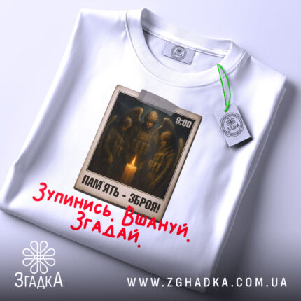 Футболка хвилина мовчання. Біла футболка, складена на поверхні, з видимим принтом на грудях. На принті зображені солдати біля свічки, текст "ПАМ'ЯТЬ - ЗБРОЯ!" та червоний напис "Зупинись. Вшануй. Згадай.". Видно етикетку з зеленим шнурком.