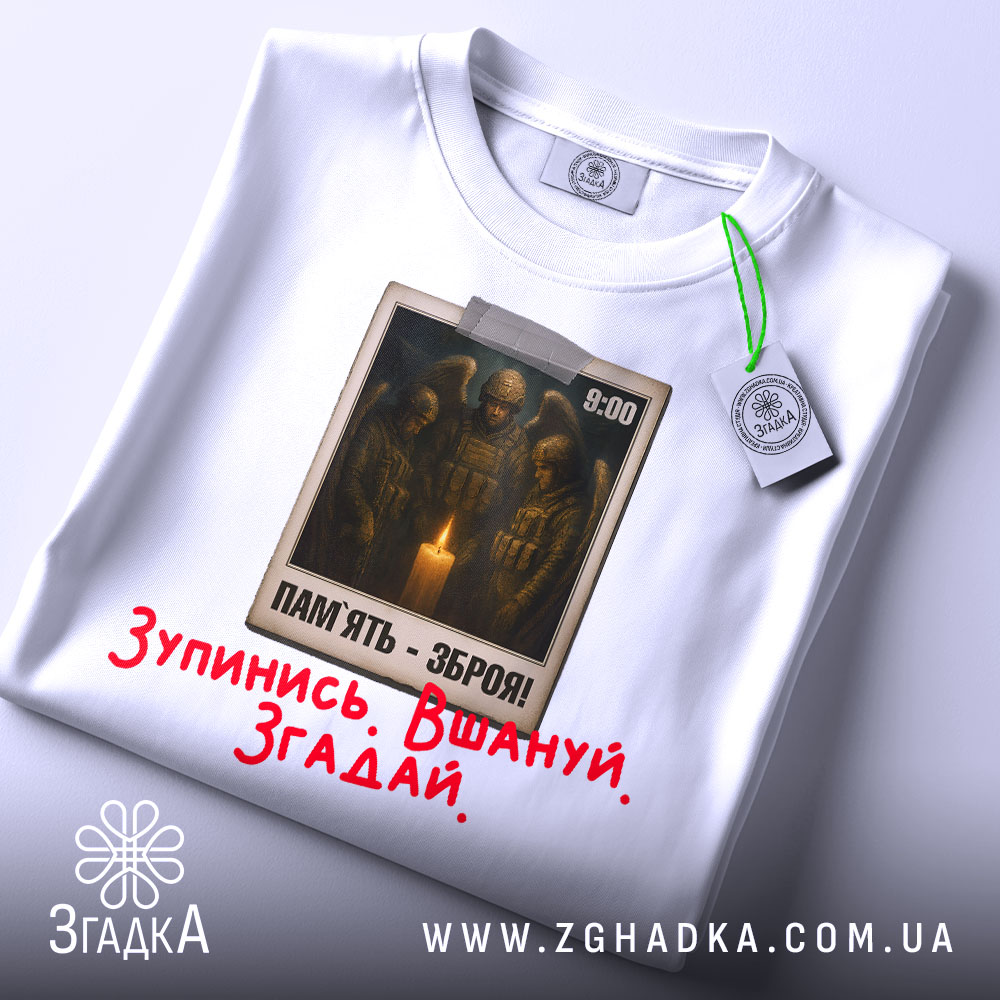 Футболка хвилина мовчання. Біла футболка, складена на поверхні, з видимим принтом на грудях. На принті зображені солдати біля свічки, текст "ПАМ'ЯТЬ - ЗБРОЯ!" та червоний напис "Зупинись. Вшануй. Згадай.". Видно етикетку з зеленим шнурком.