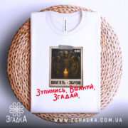 Футболка хвилина мовчання. Біла футболка, складена на плетеній основі, з видимим принтом на грудях. На принті зображені солдати біля свічки, текст "ПАМ'ЯТЬ - ЗБРОЯ!" та червоний напис "Зупинись. Вшануй. Згадай.". Видно етикетку на горловині.