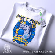 Футболка кінь боксер не жени, біла футболка складена на столі, ракурс зверху, фон світло-сірий, принт з зображенням коня в боксерських рукавичках, текст 'DON'T PUSH THE HORSES' жовтого кольору, етикетка з зеленим шнурком.