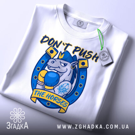 Футболка кінь боксер не жени, біла футболка складена на столі, ракурс зверху, фон світло-сірий, принт з зображенням коня в боксерських рукавичках, текст 'DON'T PUSH THE HORSES' жовтого кольору, етикетка з зеленим шнурком.