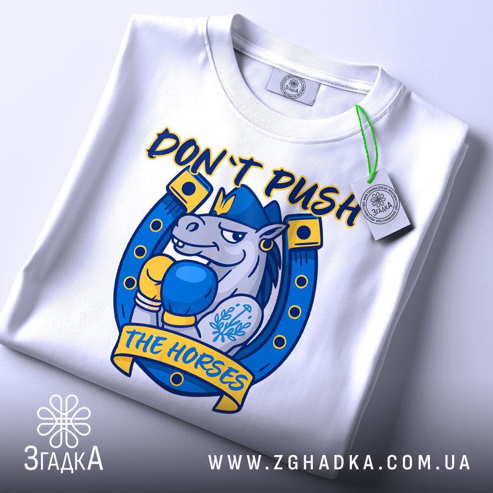 Футболка кінь боксер не жени, біла футболка складена на столі, ракурс зверху, фон світло-сірий, принт з зображенням коня в боксерських рукавичках, текст 'DON'T PUSH THE HORSES' жовтого кольору, етикетка з зеленим шнурком.
