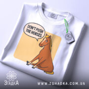 Футболка кінь don't push, біла футболка на столі, ракурс зверху, фон світло-сірий, принт з зображенням коня, що сидить, з текстом 'DON'T PUSH THE HORSES' у бульбашці, на футболці є етикетка.