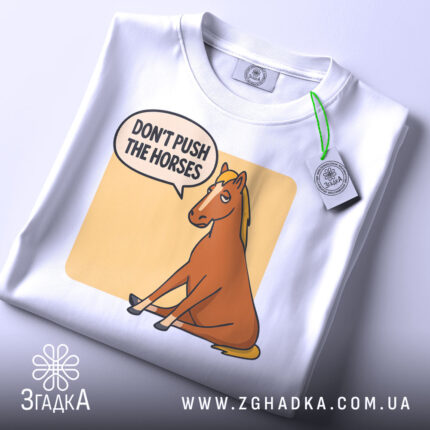 Футболка кінь don't push, біла футболка на столі, ракурс зверху, фон світло-сірий, принт з зображенням коня, що сидить, з текстом 'DON'T PUSH THE HORSES' у бульбашці, на футболці є етикетка.