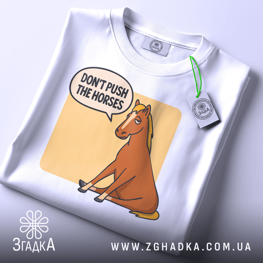 Футболка кінь don't push, біла футболка на столі, ракурс зверху, фон світло-сірий, принт з зображенням коня, що сидить, з текстом 'DON'T PUSH THE HORSES' у бульбашці, на футболці є етикетка.