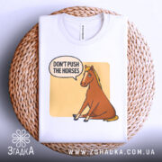 Футболка кінь don't push, біла футболка на плетеній кошику, ракурс зверху, фон світло-сірий, принт з зображенням коня, що сидить, з текстом 'DON'T PUSH THE HORSES' у бульбашці.