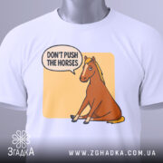 Футболка кінь don't push, біла футболка на столі, ракурс зверху, фон світло-сірий, принт з зображенням коня, що сидить, з текстом 'DON'T PUSH THE HORSES' у бульбашці, на футболці є етикетка.