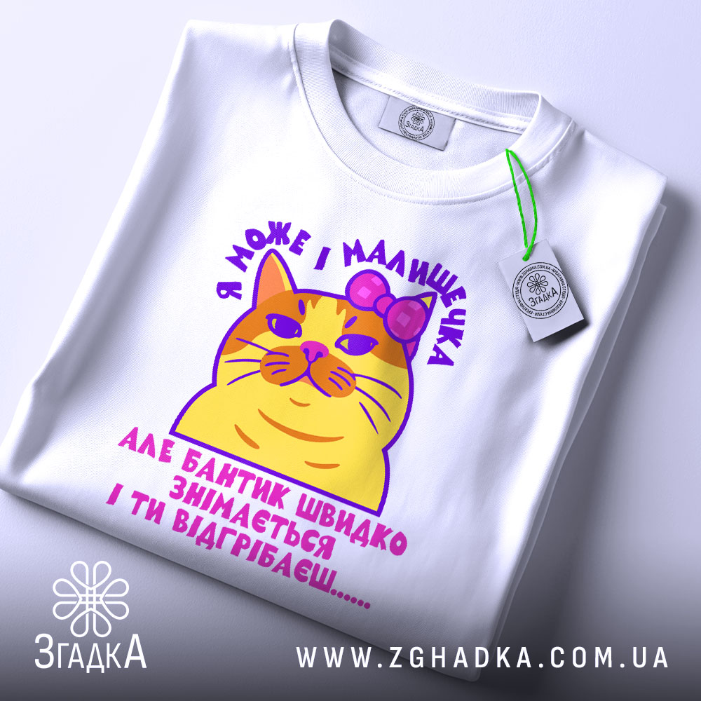 Футболка кішка малишечка, біла, зображена зверху, фон світло-сірий, принт з жовтою кішкою з рожевим бантиком та текстом, на футболці є етикетка з зеленим шнурком.