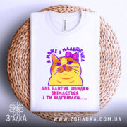 Футболка кішка малишечка, біла, зображена зверху на плетеній кошику, фон білий, принт з жовтою кішкою з рожевим бантиком та текстом на українській мові.