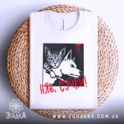 Футболка кіт бунтар, біла, складена на плетеній кошику, фон світло-сірий, принт з котом і собакою в чорному квадраті, червоний напис 'НЯВ, СУЧКИ!' з краплинами червоного.