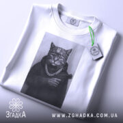 Футболка кіт гангстер стиль, біла футболка з принтом кота в гангстерському стилі, ракурс зверху, фон світло-сірий, принт зображує кота в капелюсі та з пістолетом, на шиї ланцюг, з етикеткою.