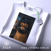 Футболка кіт геймер, біла, зображена зверху, на фоні світло-сірого градієнту, принт з котом у навушниках та масці геймера, з виразними очима, прикріплена етикетка з зеленим шнурком.