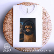 Футболка кіт геймер, біла, зображена зверху на плетеній основі, на фоні світло-сірого градієнту, принт з котом у навушниках та масці геймера, з виразними очима та деталями шерсті.